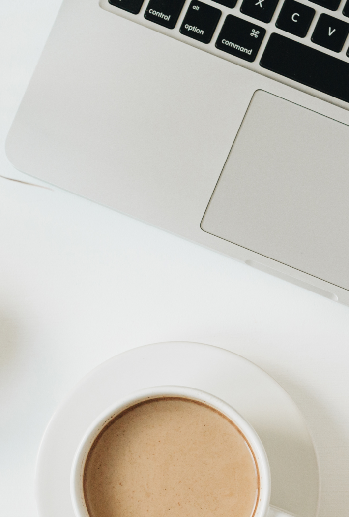 Billedet viser toppen af et hvidt skrivebord, hvor der i højre side ses det meste af et sort tastatur på en grå Macbook. Nederst ses en kop kaffe med mælk, og i venstre side ses der nederst en lille hvid skål med gyldne papirklips, og øverst et par lange spidse stængler af en eukalyptus-grøn grøn plante