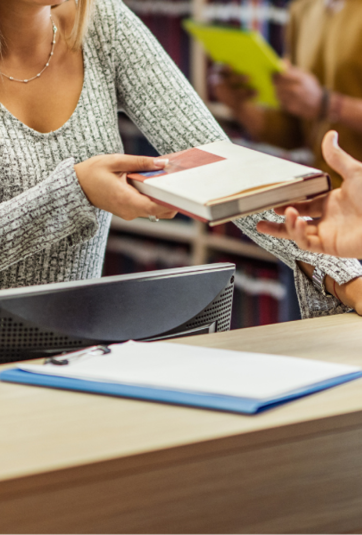 Billedet viser en kvinde giver en bog til en anden person ved et bord i et bibliotek.
