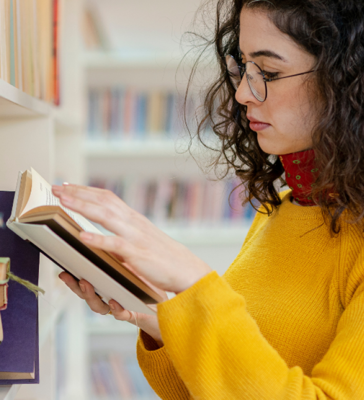 En kvinde med skulerlangt mørkt krøllet hår og briller, iført en gul sweater og rødt tørklæde om halsen. Står og kigger i en bog ved en lys bogreol
