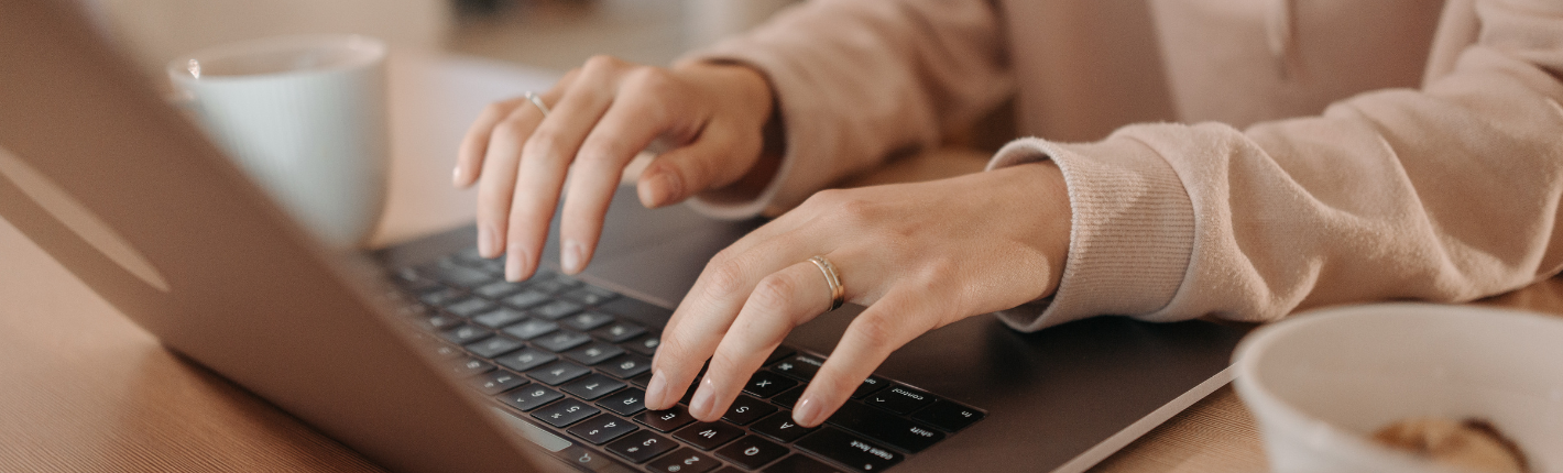 Et nærbillede af en persons hænder, der skriver på et bærbart tastatur. Personen bærer en guldring på venstre hånd, som hviler oven på tasterne. Selve den bærbare computer er sort med sølvfarvede detaljer, og dens skærm viser en række ikoner. En skål fyldt med forskellige genstande sidder til højre for den bærbare computer, mens en glaskop kan ses i baggrunden i nederste venstre hjørne.